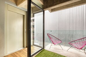 Un balcon moderne avec une porte vitrée, deux chaises rouges à structure métallique et une petite portion de gazon synthétique.