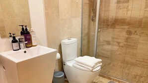 Ein Badezimmer mit Waschbecken, Toilette und Dusche. Auf dem Waschbecken stehen Pflegeprodukte, und ein Mülleimer ist sichtbar.