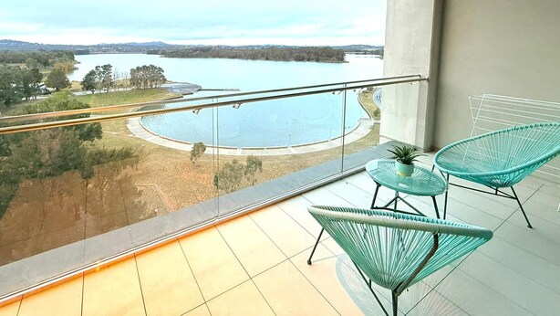 Uma varanda com vista para um lago e uma ponte, mobiliada com duas cadeiras verdes e uma mesa pequena.