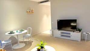 Uma sala de estar moderna com uma mesa redonda branca, um sofá, uma TV sobre um suporte branco e uma pequena mesa lateral branca.