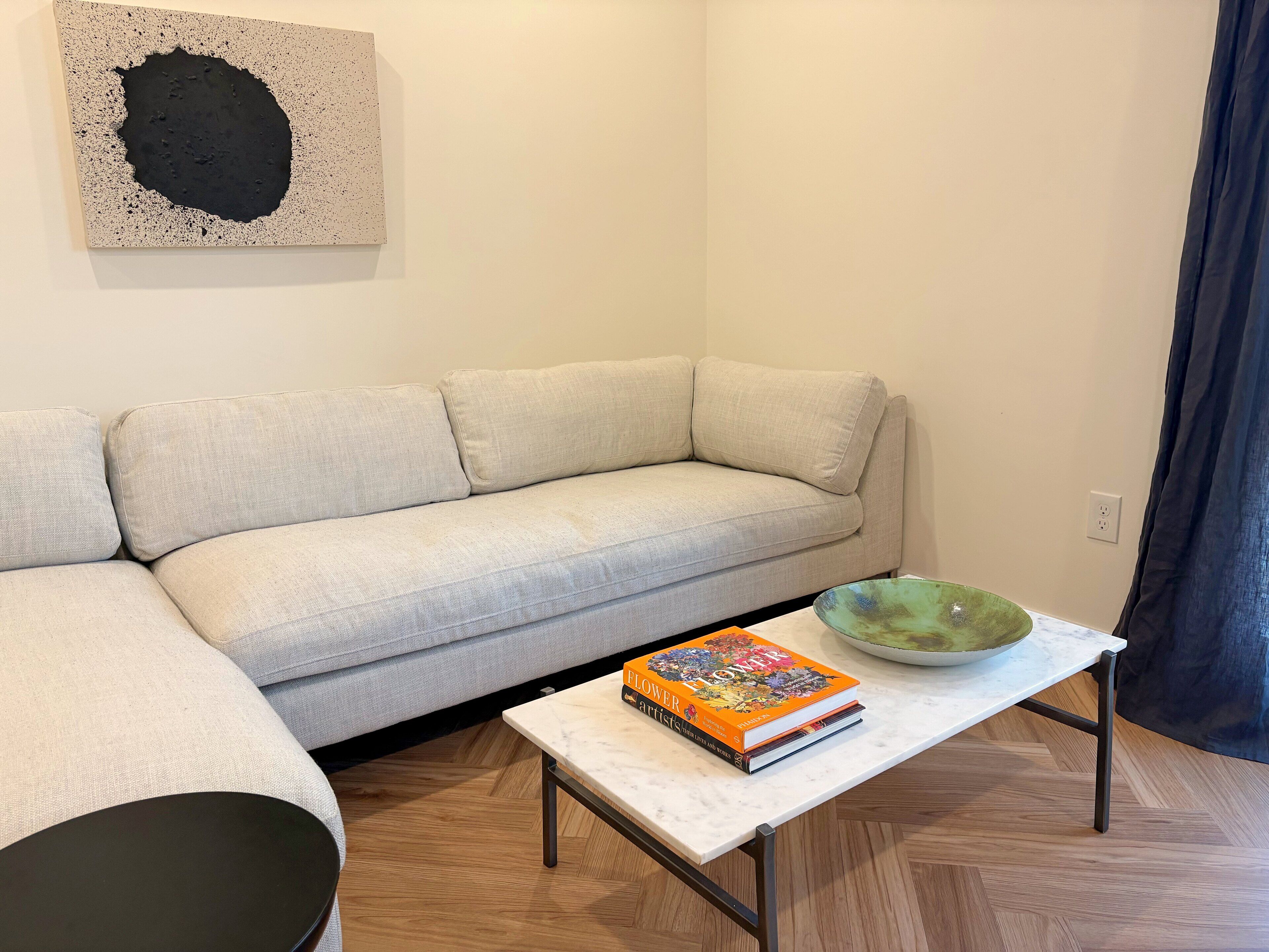 A living room with a beige sofa, a white coffee table with a book and a green bowl, and a black abstract painting on the wall.
