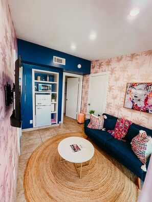 Una sala de estar con un sofá azul, una mesa de centro blanca y un tapete estampado. Hay una zona de cocina con microondas y refrigerador.
