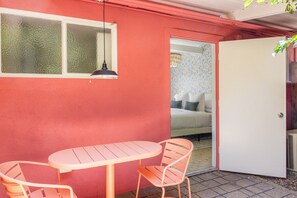 Una habitación con pared roja, puerta blanca, cama con ropa de cama estampada y una pequeña mesa con dos sillas.