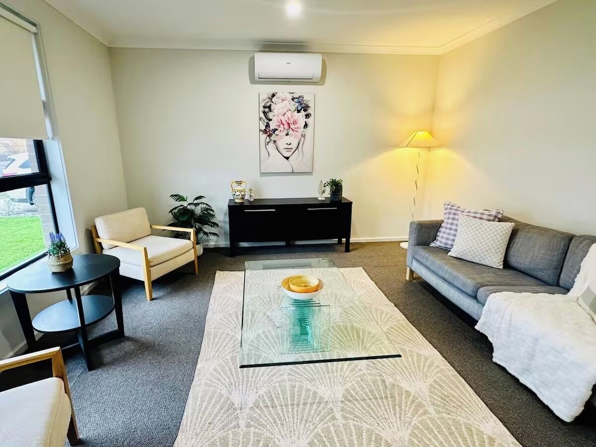 A modern living room with a glass coffee table, a grey sofa, a beige armchair, a black side table, and a decorative wall piece.