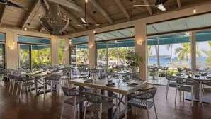 Un comedor amplio con varias mesas y sillas, ventanas grandes que ofrecen vista al mar, y un techo alto con vigas a la vista.