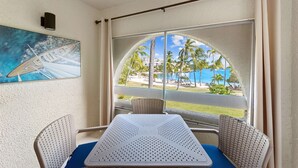 Una habitación con una mesa y sillas, un cuadro de un barco en la pared, y vistas a una playa y palmeras a través de amplios ventanales.