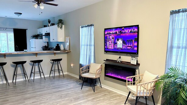 Una sala de estar moderna con televisor, taburetes de bar, chimenea y una zona de cocina con refrigerador y armarios.