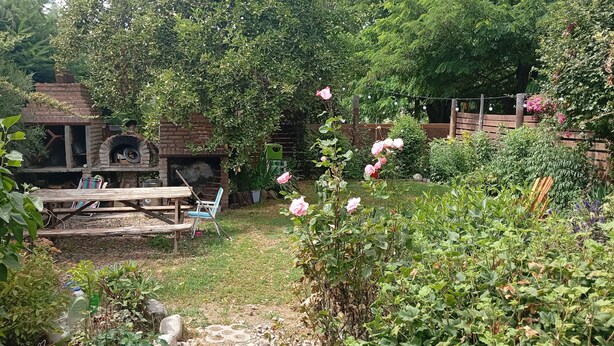 A rustic outdoor area with a brick oven, wooden table, and chairs, surrounded by lush greenery and flowering plants. - Los Colibríes duplex for rent (pet friendly) (Chubut)