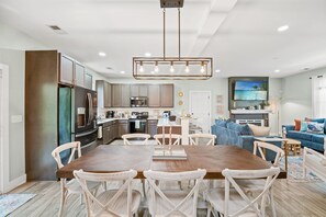 A modern kitchen with stainless steel appliances, a dining area with a wooden table and white chairs, and a living area with a blue sofa and a TV. - Sleeps 10 - Private Pool - Close to Folly & Kiawah (Johns Island)