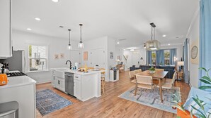 A modern kitchen with white cabinets, a central island, and pendant lighting. There is a dining area with a table and chairs, and a living room with a sofa and a coffee table. - Close 2 DT & Beaches | 4 BR 10 Guests | Built 2025 (Charleston)