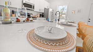 A modern kitchen with white cabinets, a central island with a glass top, and a dining area with a woven placemat. - Close 2 DT & Beaches | 4 BR 10 Guests | Built 2025 (Charleston)