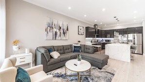Ein modernes Wohnzimmer mit einem grauen Ecksofa, einem runden Couchtisch und einem weißen Beistelltisch. Im Hintergrund ist eine Küche mit dunklen Schränken und Edelstahlgeräten zu sehen.