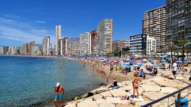 Una platja amb gent prenent el sol i banyant-se, un litoral rocos i edificis alts al fons.