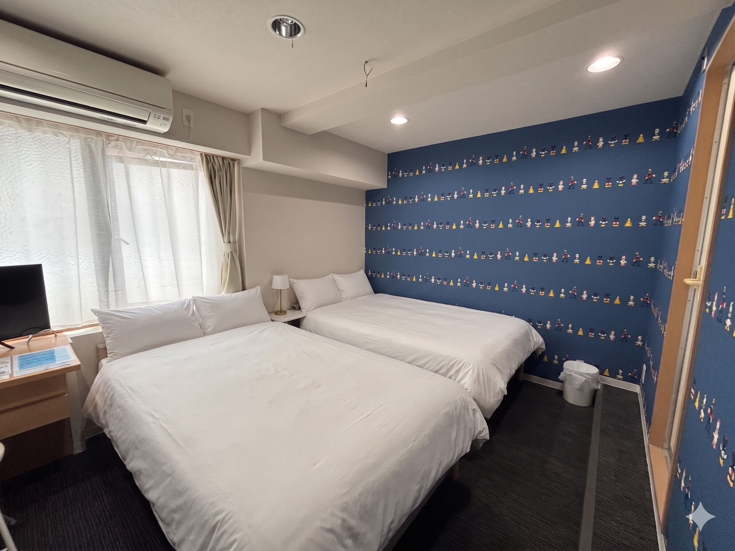 Ein Hotelzimmer mit zwei Betten, einem Schreibtisch mit Computer, einer Klimaanlage und einer blauen Wand mit Zeichentrickfiguren.