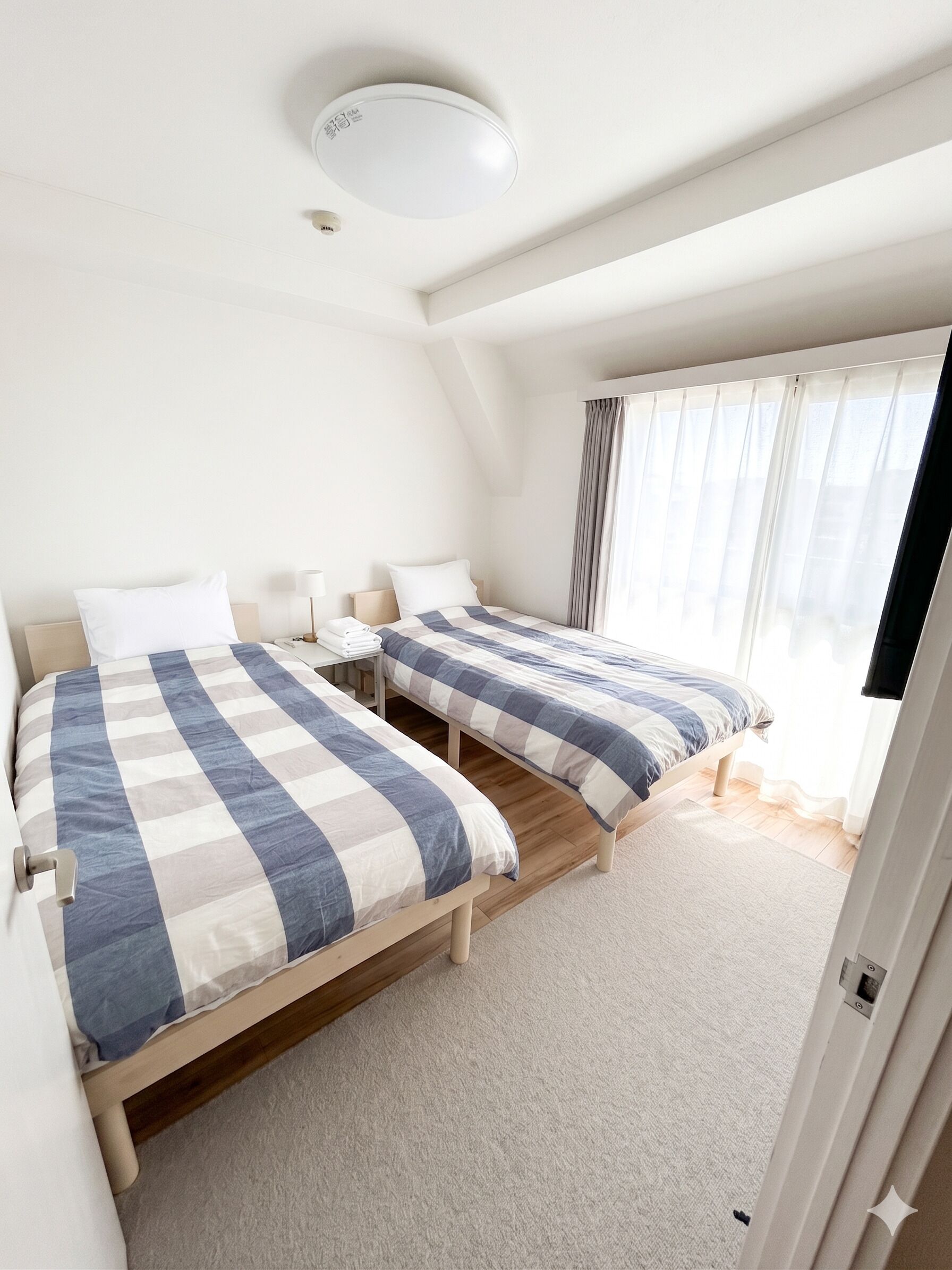 Una habitación con dos camas, una lámpara en el techo, una ventana con cortinas y una puerta.