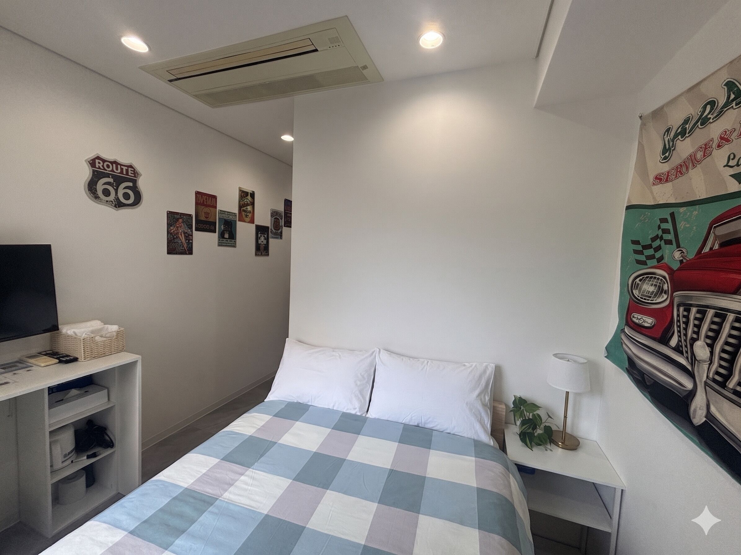 A bedroom with a bed, a TV, a shelf with items, a wall with Route 66 posters, and a vintage car poster.