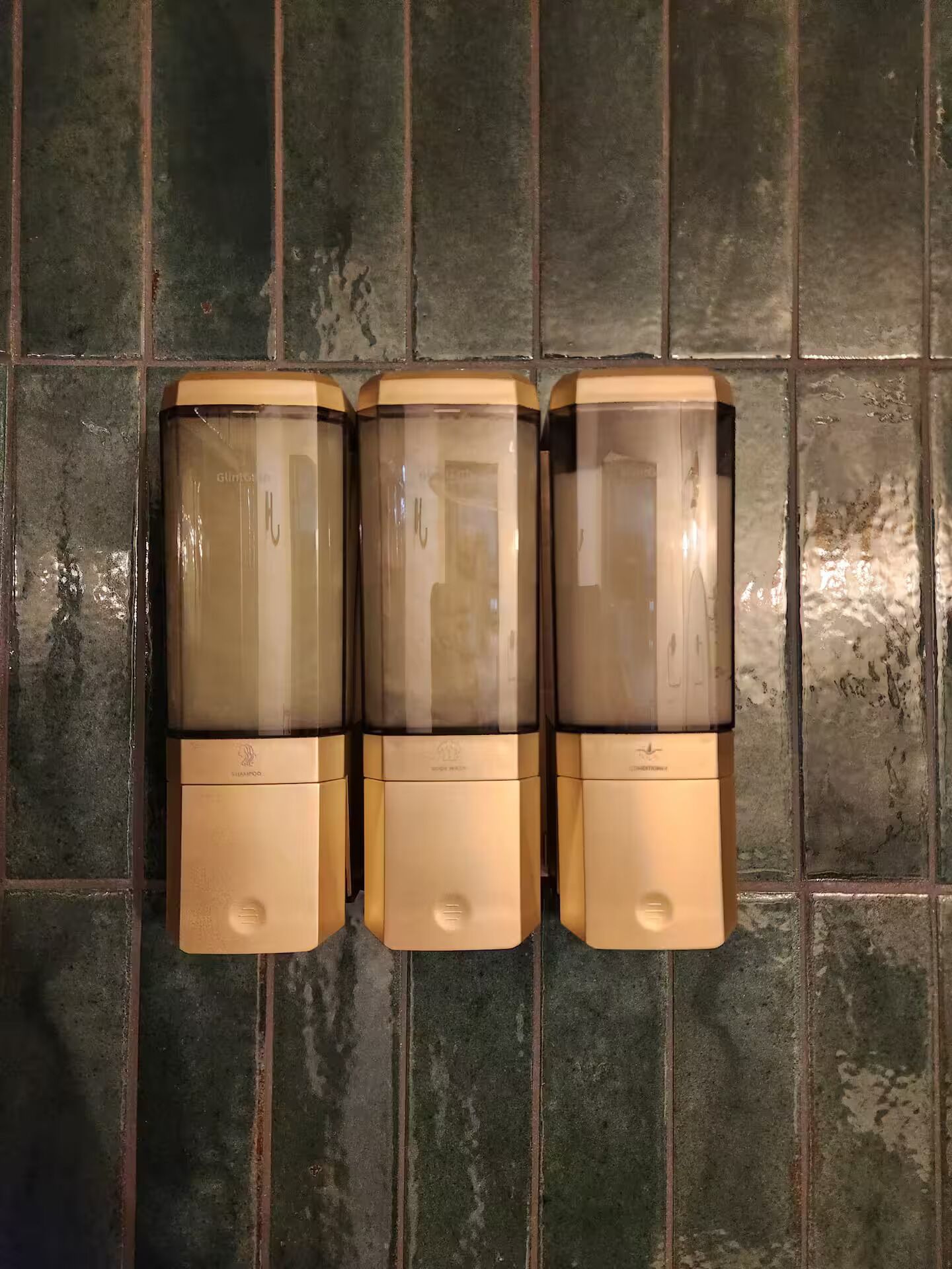 Drei Shampoodispenser an einer gefliesten Wand.