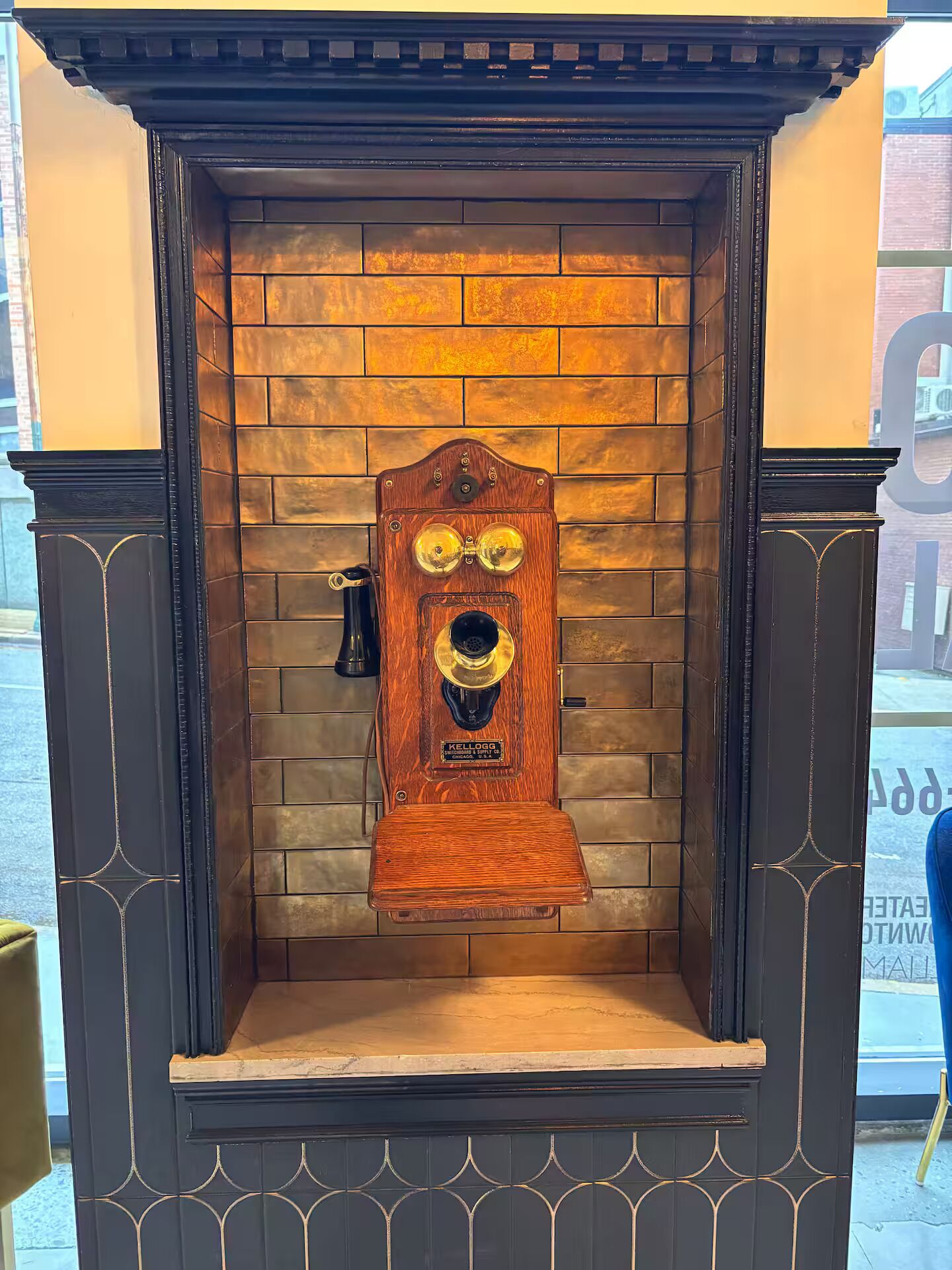 Eine Telefonzelle im Vintage-Stil mit Holzaußenverkleidung und schwarzem Fuß, platziert in einer Ecke mit Backsteinwänden und schwarzem Zierstreifen.