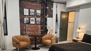 Hotellihuone, jossa on yksi seinä koristeltu tiilillä, kaksi nojatuolia, pyöreä pöytä, mustalla päällisellä varustettu sänky ja kattokruunu.