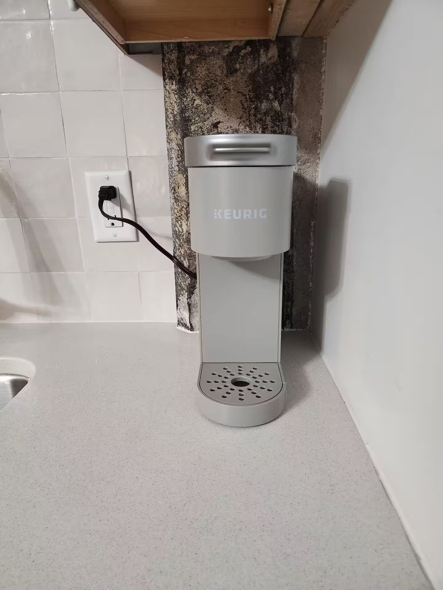 Uma cafeteira Keurig sobre uma bancada de cozinha com um painel de azulejos brancos e um armário de madeira acima.
