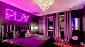 Ein modernes Schlafzimmer mit einem großen Bett, einem Neon-Schild mit der Aufschrift 'PLAY', einer Kristallleuchtre und Blick auf die beleuchtete Stadt bei Nacht.
