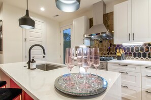 A modern kitchen with white cabinetry, a stainless steel range hood, and a glass-topped island with wine glasses. - Designer Home - 10beds! Rooftop with Hot Tub! Close 2 DT! (Nashville)