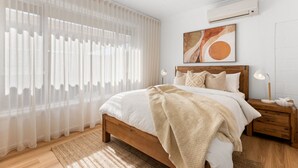 A bedroom with a wooden bed, white bedding, a large window with sheer curtains, a ceiling fan, an air conditioning unit, and a modern abstract painting on the wall. - The Driftwood - Explore the Beach Life at Henley (Henley Beach)