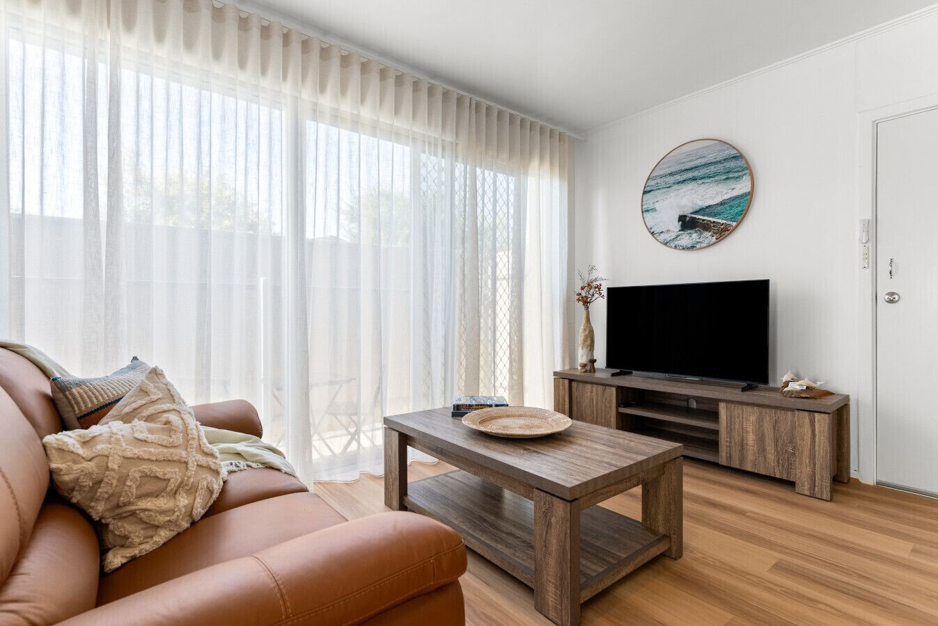 A modern living room with a leather sofa, a wooden coffee table, a flat-screen TV, and a large window with sheer curtains.
