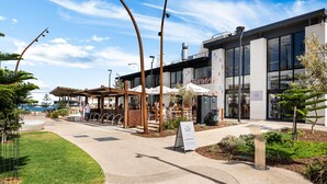 An outdoor cafe area with tables and chairs, a signboard, and a modern building with large windows. - The Driftwood - Explore the Beach Life at Henley (Henley Beach)