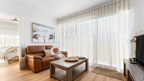 A modern living room with a brown leather sofa, a wooden coffee table, and a large window with sheer curtains. - The Driftwood - Explore the Beach Life at Henley (Henley Beach)