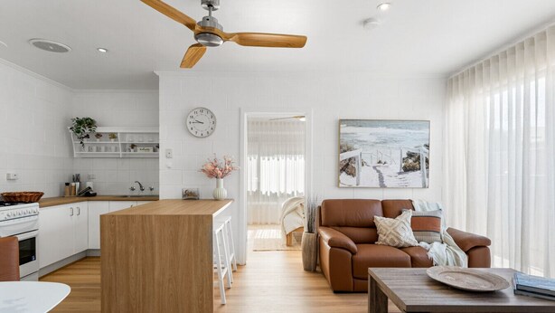 A modern kitchen with white cabinets, a wooden island, and a dining table. There is a living area with a brown leather sofa and a coffee table. A ceiling fan is visible, and a painting hangs on the wall. - The Driftwood - Explore the Beach Life at Henley (Henley Beach)