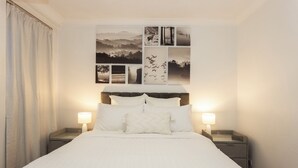 Cama bem arrumada com roupa de cama branca, duas mesas de cabeceira com abajures e uma série de fotografias a preto e branco na parede acima da cama.