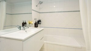 Casa de banho com uma pia branca, uma banheira e um chuveiro. Há produtos de higiene na pia e um porta-toalhas em cima.