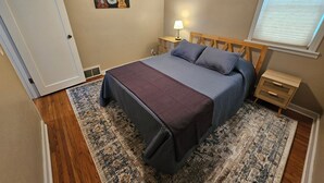 A bedroom with a bed, a nightstand, a lamp, a window with blinds, and a door.