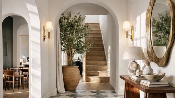 Um corredor com chão xadrez, portas arqueadas, escada e um console com espelho e objetos decorativos.