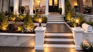 Ein zweistöckiges Haus mit einer Veranda, Treppen zum Eingang und einem gut beleuchteten Garten.