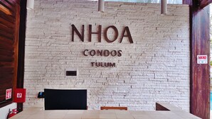 Vastaanottoalue, jossa on valkoinen tiiliseinä, digitaalinen näyttö ja kyltti, jossa lukee: ”NHOA CONDOS TULUM”.