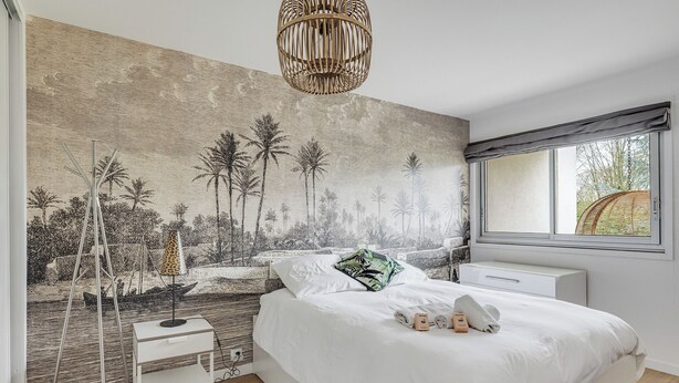 Ein Schlafzimmer mit einer großen Wandmalerei einer tropischen Szene, einem weißen Bett mit Kissen, einem Nachttisch und einem Fenster mit Blick auf Grünflächen.