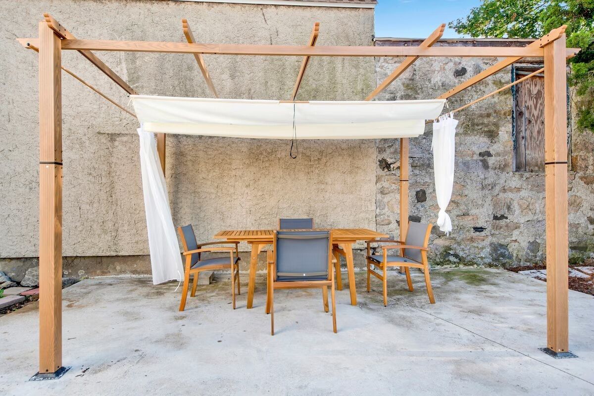Una pérgola de madera con toldo blanco, una mesa redonda y seis sillas en un entorno al aire libre.