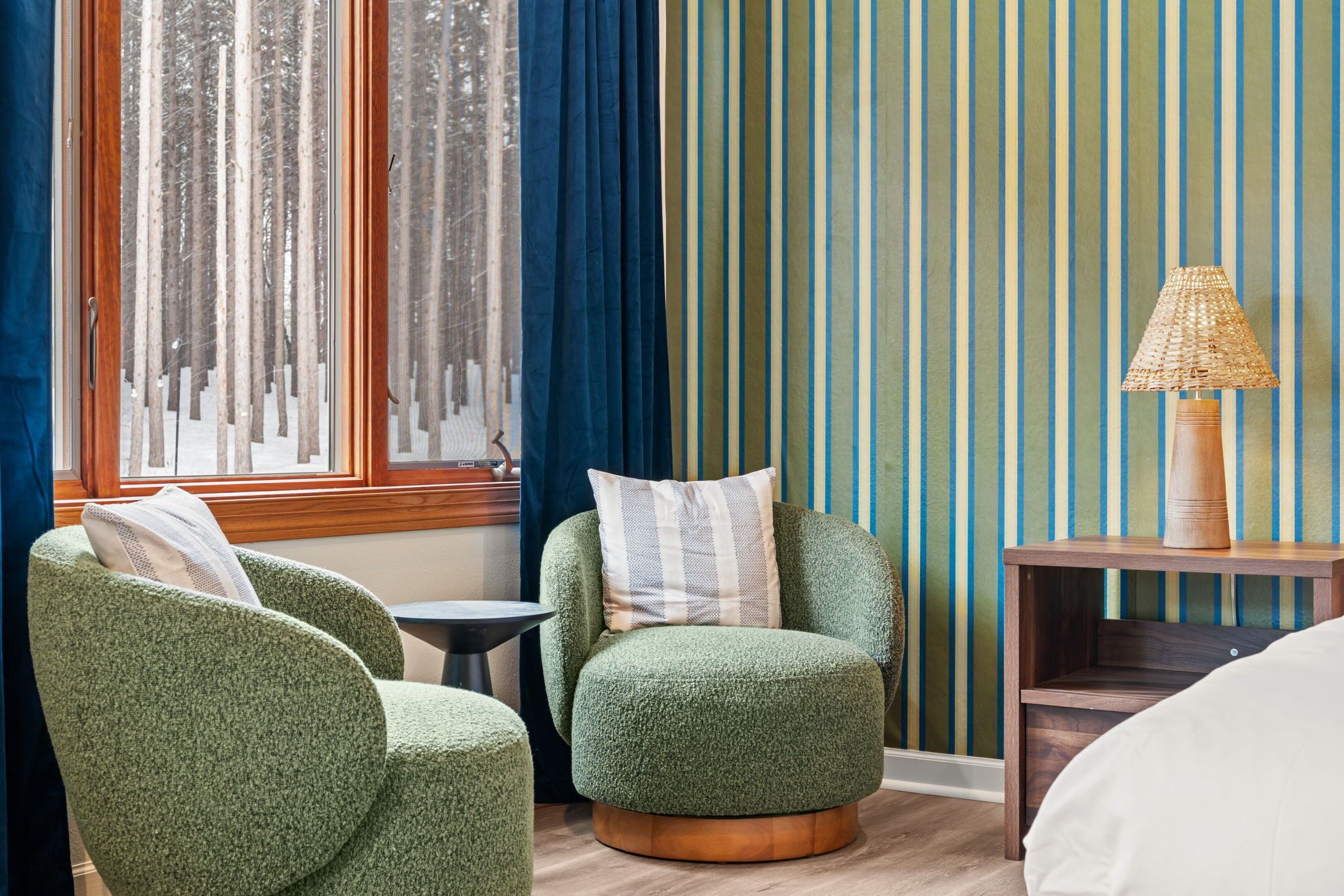 Une pièce comprenant deux fauteuils verts, une petite table ronde, une table d’appoint en bois avec une lampe, et une fenêtre avec des rideaux.