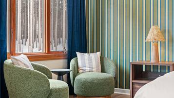 Une pièce comprenant deux fauteuils verts, une petite table ronde, une table d’appoint en bois avec une lampe, et une fenêtre avec des rideaux.