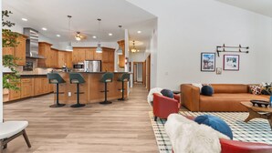 A modern kitchen with wooden cabinets, a central island with bar stools, and a dining area with a red sofa and patterned rug. - The Powder Haus: Hot Tub, Sauna, Fire Pit, Golfsim (Wisconsin Dells)
