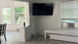 Uma sala de estar espaçosa com uma televisão de tela grande fixada na parede, um rack branco e uma janela grande com persianas.