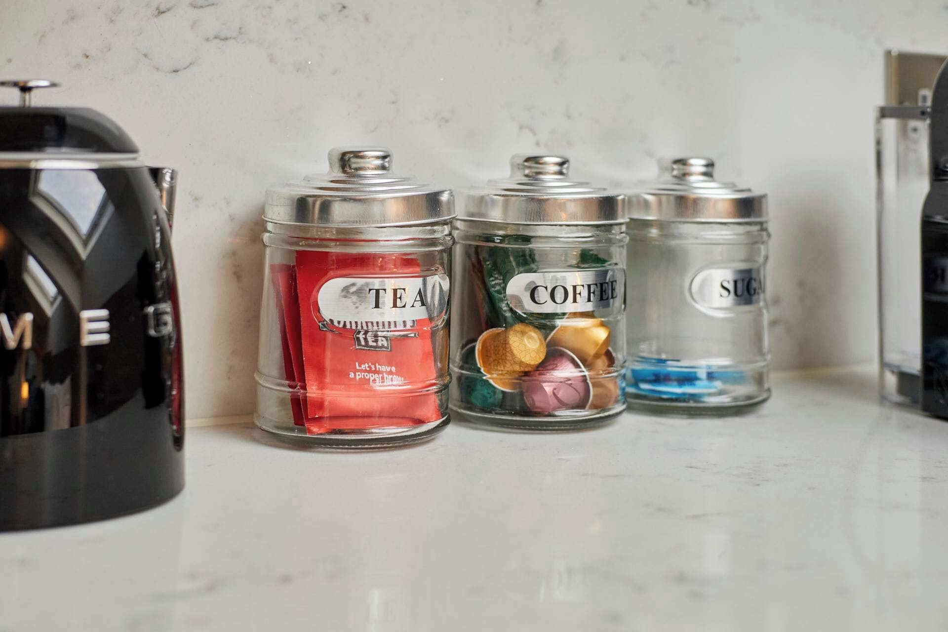 Three glass containers with metal lids labeled "TEA," "COFFEE," and "SUGAR" on a marble countertop.