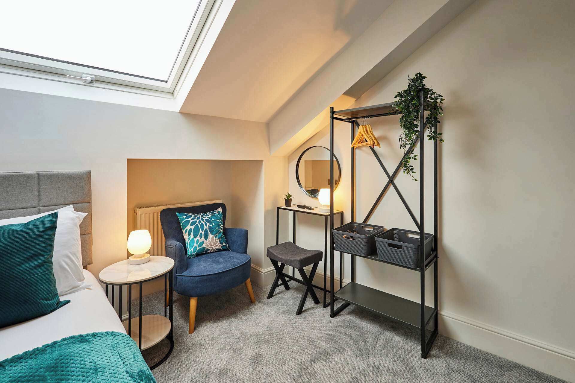 Habitación de hotel compacta con cama, una pequeña mesa redonda con lámpara, una silla azul, un escritorio con espejo y un estante metálico negro con cestas.