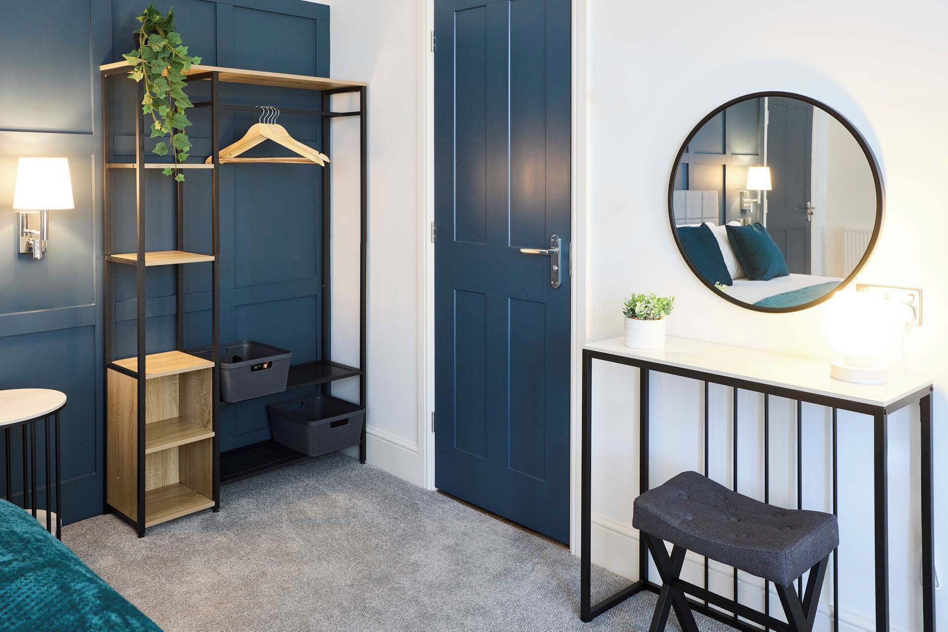 A room with a dark blue door, a round mirror, a small stool, and a shelving unit with a plant and a hanger.