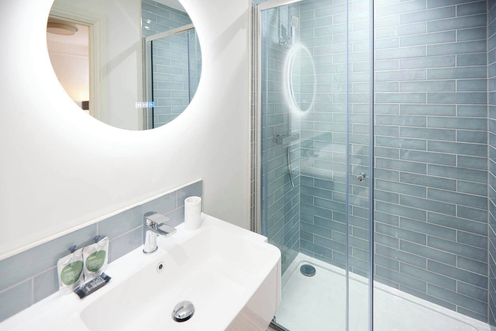 A bathroom with a white sink, a mirror, and a glass shower enclosure with blue tiles.