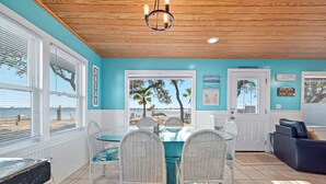 A dining area with a table and chairs, a view of the beach through large windows, and a door with a sign. - Bama Breeze | Little Lagoon w/ Fishing Dock | Dog Friendly | My Beach Getaways (Gulf Shores)