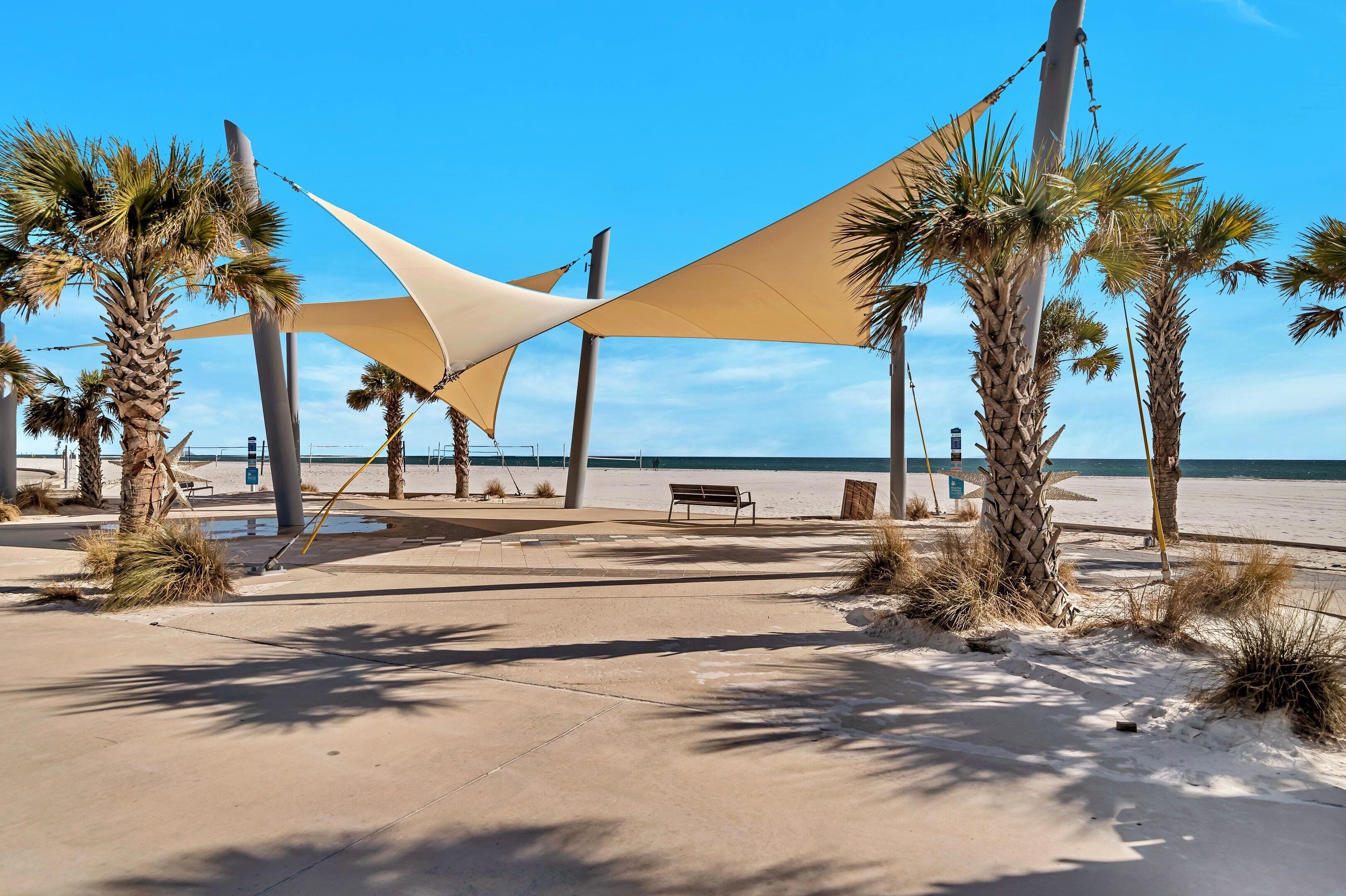 A beachside walkway with palm trees, a bench, and a large shade structure.