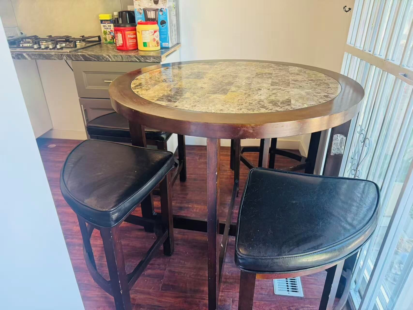 Una mesa de comedor redonda con dos sillas negras, una cocina con estufa a gas y una ventana con persianas verticales.